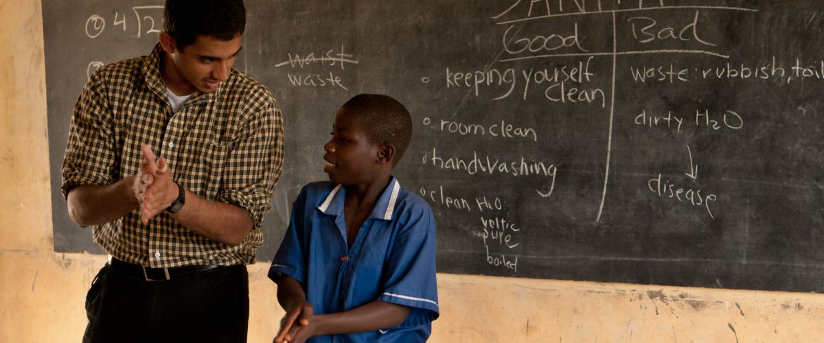 teacher and a young student stand in front of a blackboard, practicing handwashing gestures